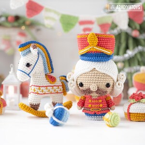 Op de afbeelding: Gehaakte kerstversieringen, waaronder een wit schommelpaard met blauwe en gele accenten, een notenkrakersoldaat en ingepakte cadeaus. De speelgoedjes zijn handgemaakt met kleurrijk garen en staan tegen een feestelijke achtergrond met een kerstboom.