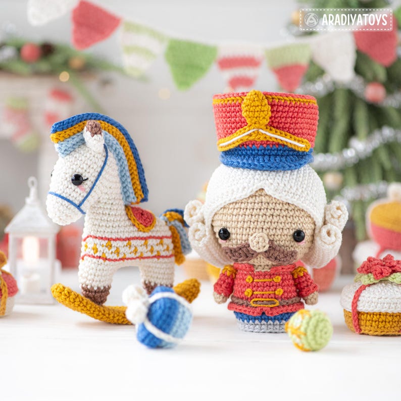 Op de afbeelding: Gehaakte kerstversieringen, waaronder een wit schommelpaard met blauwe en gele accenten, een notenkrakersoldaat en ingepakte cadeaus. De speelgoedjes zijn handgemaakt met kleurrijk garen en staan tegen een feestelijke achtergrond met een kerstboom.