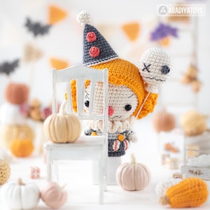May include: A crocheted clown doll with orange hair, a dark gray cone hat with red pom-poms, and a ruffled collar. The doll holds a white balloon with a stitched face and is partially obscured by a white chair. The text ARADIYATOYS is visible.