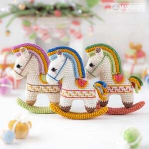 May include: Three handmade crochet rocking horses in cream, each with a colorful mane and saddle. The horses have a detailed design on their bodies and are set on a wooden base. The background includes Christmas decorations.