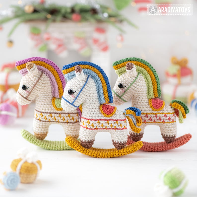 May include: Three handmade crochet rocking horses in cream, each with a colorful mane and saddle. The horses have a detailed design on their bodies and are set on a wooden base. The background includes Christmas decorations.