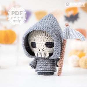 Puede incluir: Muñeco de la Parca tejido a ganchillo con cara de calavera, con una túnica con capucha gris y una guadaña. La calavera es de color beige claro con cuencas de los ojos y boca de color gris oscuro. El texto "PDF pattern only" está en un círculo blanco.