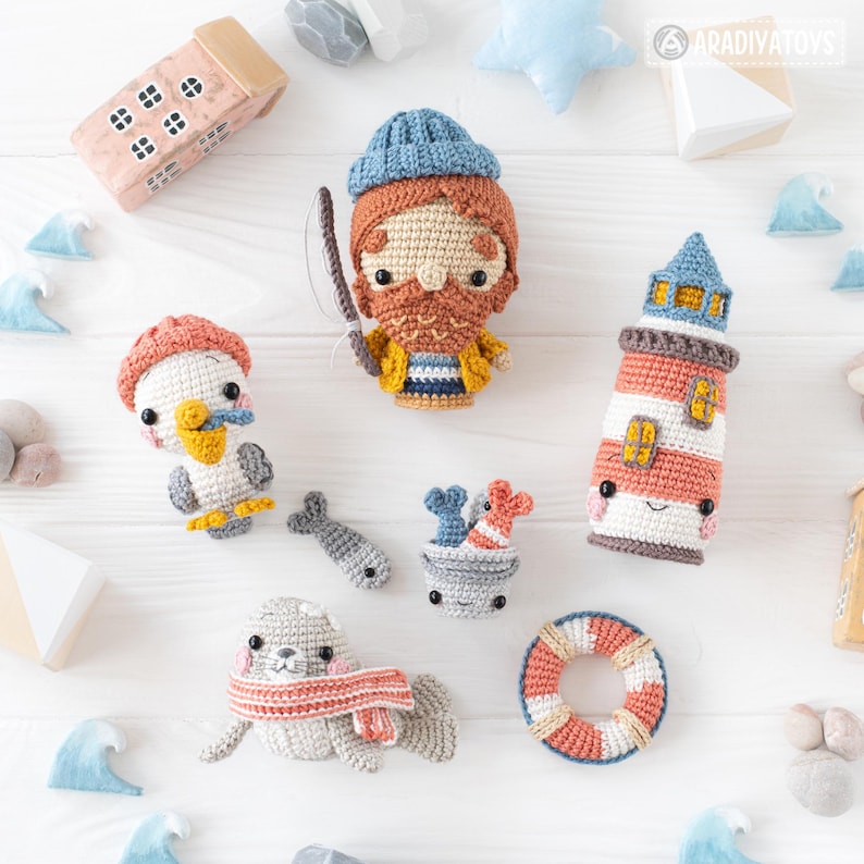 Może przedstawiać: Zestaw pluszowych zabawek amigurumi wykonanych na szydełku w morskim stylu. Zestaw zawiera rybaka, latarnię morską, mewę, fokę, wiaderko z rybami i koło ratunkowe. Zabawki są wykonane z miękkiej przędzy i mają urocze buźki.