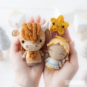 May include: A crocheted nativity scene featuring a brown and white highland cow, a blonde angel with a halo, and a yellow star.