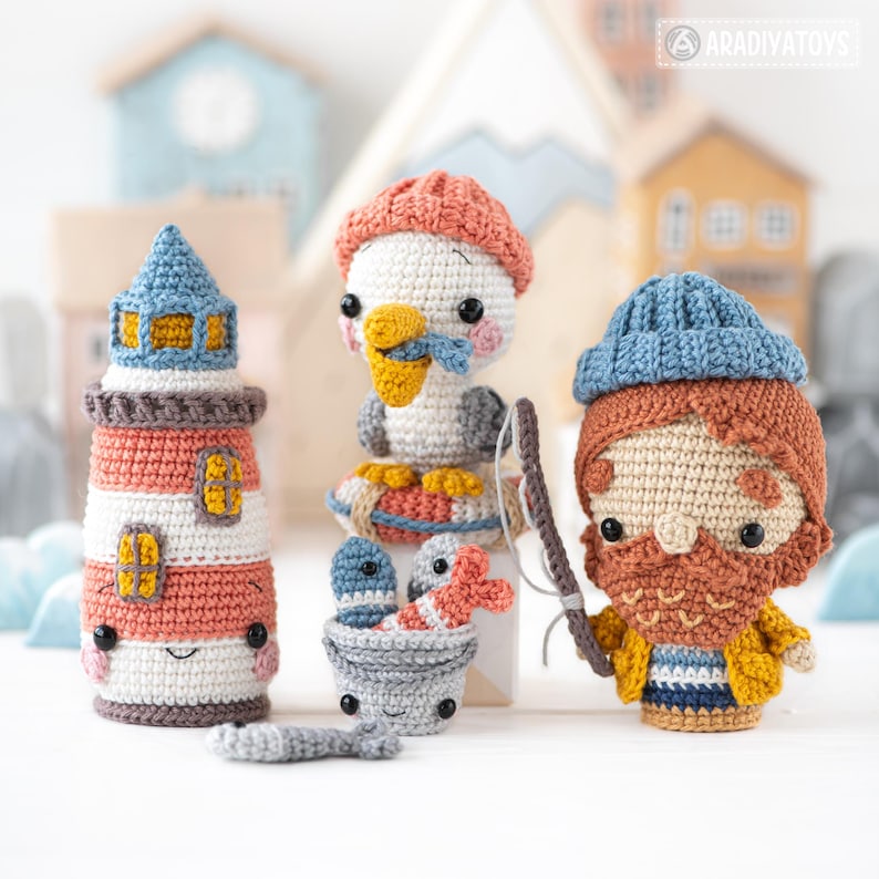Może przedstawiać: Zestaw trzech pluszak&oacute;w amigurumi wykonanych na szydełku: latarnia morska w czerwono-białe pasy, mewa z czerwoną kołem ratunkowym i rybak w niebieskim kapeluszu z wędką. Wszystkie pluszaki uśmiechają się i mają duże oczy.