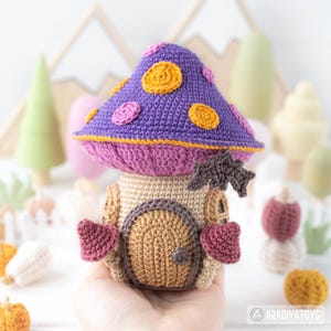 May include: A handmade crochet mushroom house with a purple cap adorned with orange and pink circles. The house features a tan body with a brown door and heart-shaped accents. A small, brown bat-shaped embellishment is attached to the house.
