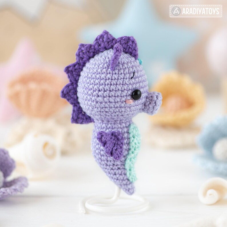 May include: A handcrafted crochet seahorse in shades of purple and mint green. The amigurumi seahorse has a detailed fin, a spiky dorsal fin, and a sweet expression. The handmade toy is displayed on a white stand, with other sea-themed decorations in the background.