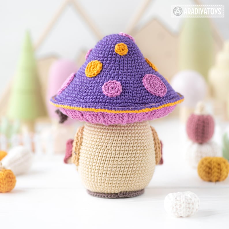 May include: A handmade crochet mushroom toy. The mushroom cap is purple with yellow and pink polka dots, and a pink and yellow trim. The stem is beige with brown accents. The toy is set against a soft, blurred background with other crochet items.