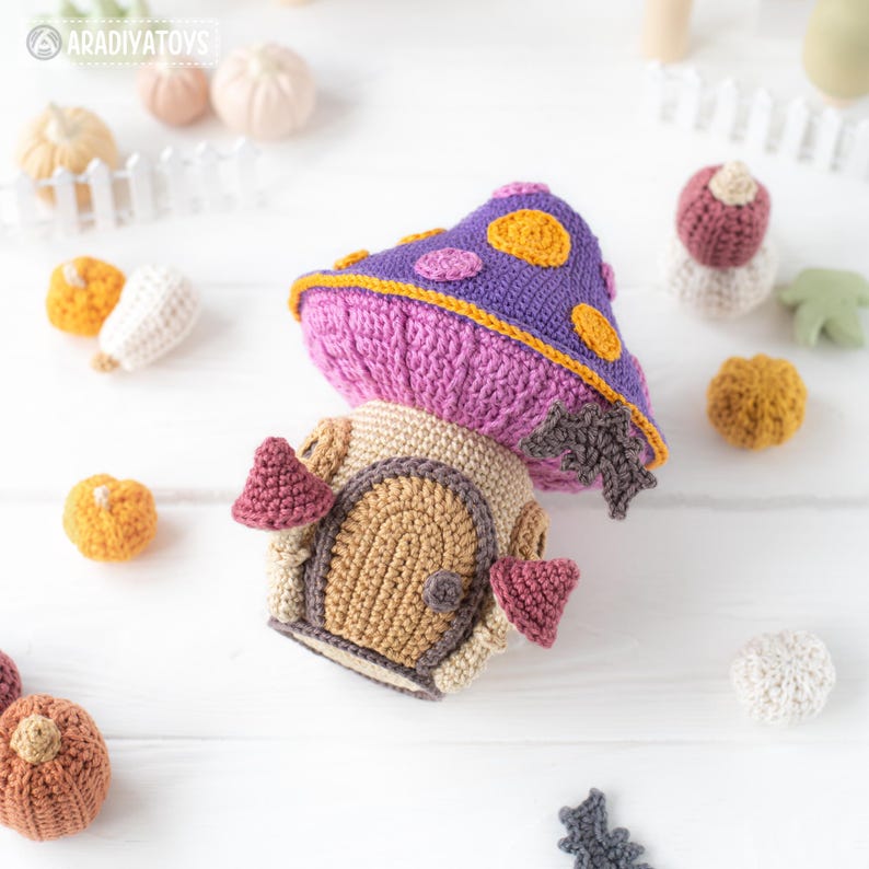 May include: A crocheted mushroom house with a purple cap featuring yellow polka dots and a pink stem. The house has a tan door and pink accents. Other crocheted items, including pumpkins and leaves, are scattered around.