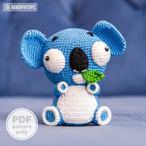 May include: A crocheted blue and white koala holding a green leaf. The koala has large, white eyes and a black nose. The koala is sitting on a blue surface. PDF pattern only.