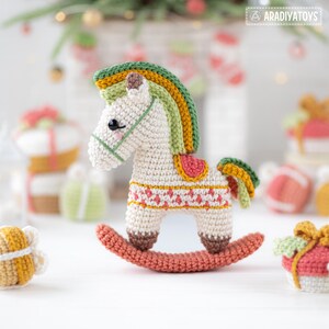 May include: A crocheted rocking horse in cream, with a colorful mane and tail in shades of green, yellow, and orange. The horse has a red saddle and is on a coral-colored rocker. The background includes other crocheted gifts.
