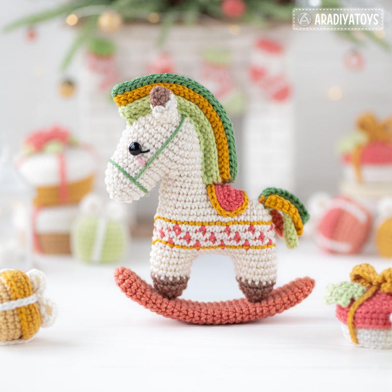 May include: A crocheted rocking horse in cream, with a colorful mane and tail in shades of green, yellow, and orange. The horse has a red saddle and is on a coral-colored rocker. The background includes other crocheted gifts.