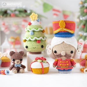 Op de afbeelding: Een verzameling handgemaakte gehaakte kerstversieringen. Bevat een groene kerstboom met een gele ster, een bruine teddybeer met een blauwe strik, een cadeauverpakking en een notenkrakersoldaat. De ornamenten zijn kleurrijk en hebben vriendelijke gezichten.