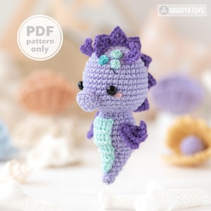 Peut inclure: Un hippocampe crocheté aux couleurs violettes et turquoise. L'hippocampe a des yeux noirs, des joues roses et une nageoire dorsale épineuse. Le texte "PDF pattern only" est dans un cercle blanc. Objet fait main.