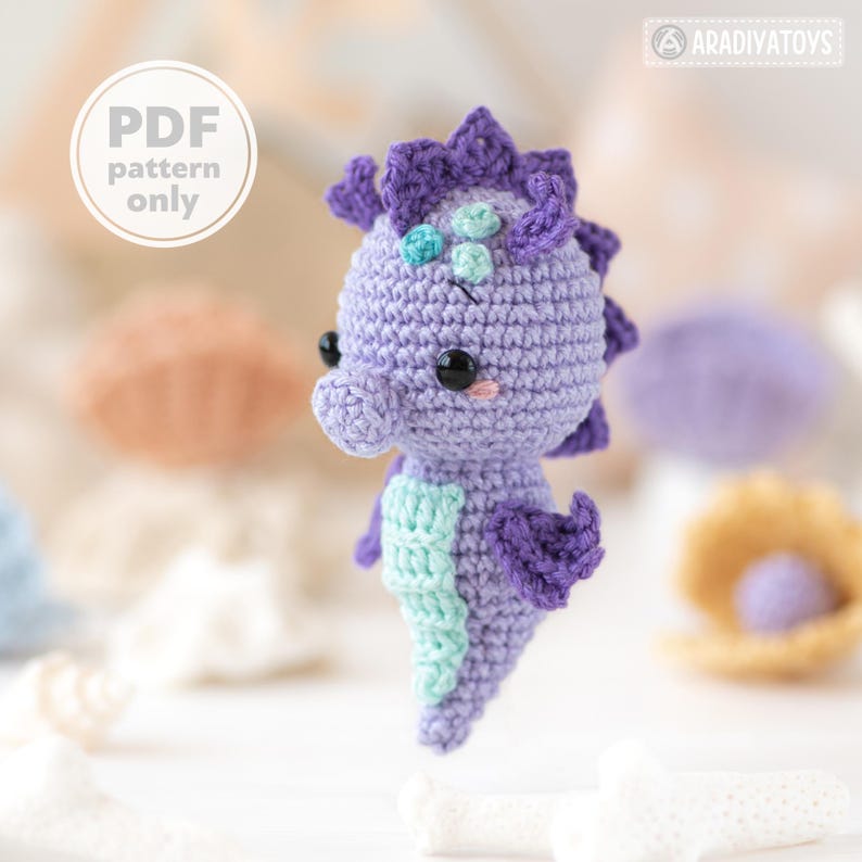 May include: A crocheted seahorse in shades of purple and teal. The seahorse has black eyes, a light pink blush, and a spiky dorsal fin. The text "PDF pattern only" is in a white circle in the upper left corner. The seahorse is a handmade item.