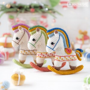 May include: Three handmade crochet rocking horses in a row. Each horse has a white body, colorful mane, and a patterned saddle blanket. The rocking bases are in shades of green, coral, and gold. The background is blurred with holiday decorations.