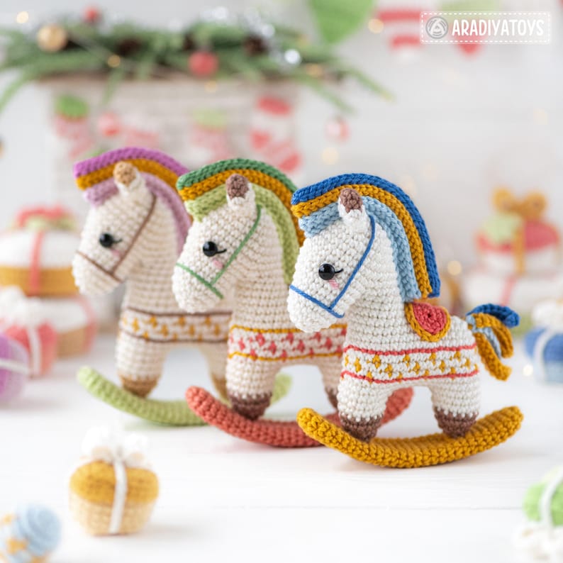 May include: Three handmade crochet rocking horses in a row. Each horse has a white body, colorful mane, and a patterned saddle blanket. The rocking bases are in shades of green, coral, and gold. The background is blurred with holiday decorations.