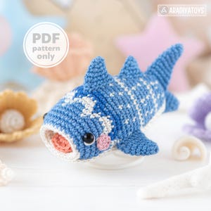 May include: A handmade blue and white crochet whale shark toy with a pink mouth and black eyes. The toy has white spots and fins. The text "PDF pattern only" is in a white circle in the upper left corner. The toy is on a white surface.