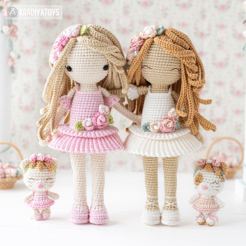May include: Two handmade crochet dolls with long hair and floral headbands, one in pink and one in white, stand side-by-side. Two smaller matching dolls are also present. The dolls are wearing dresses and have closed eyes. The brand name "ARADIYATOYS" is visible.