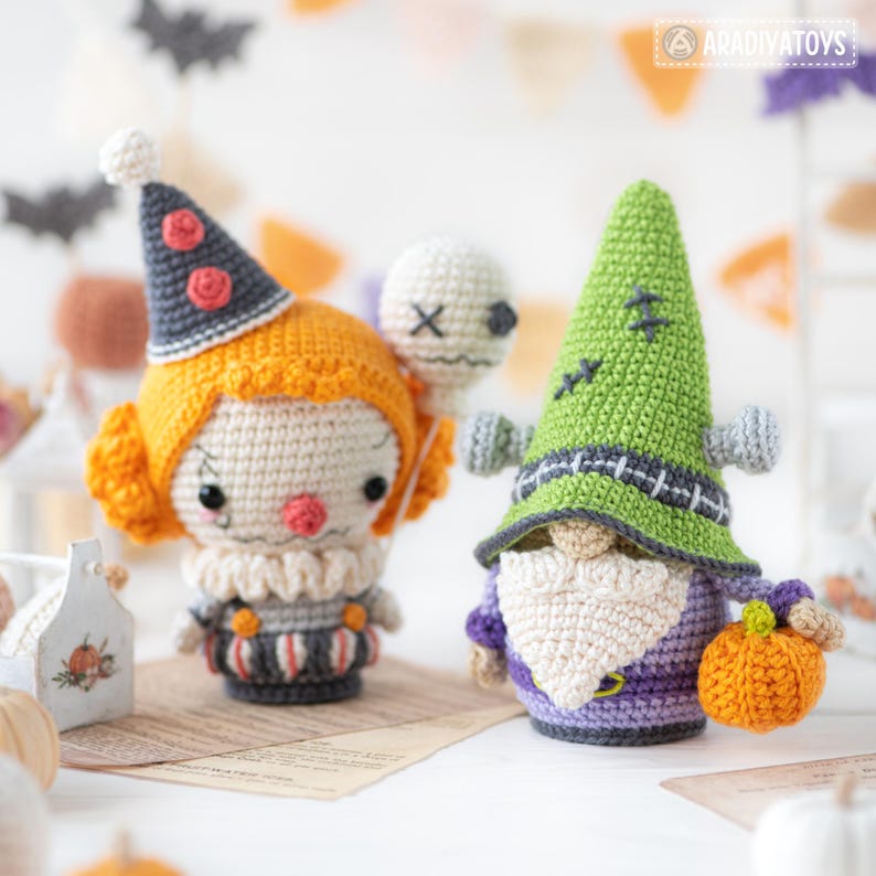 May include: Two crochet dolls: one is a clown with orange hair, a gray hat with red accents, and a white ruff collar holding a white skull balloon; the other is a green-hatted creature with a white beard, purple body, and an orange pumpkin.