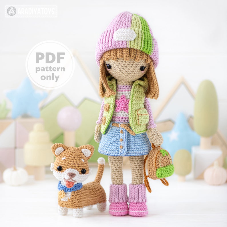 K&ouml;nnte beinhalten: H&auml;kelmuster f&uuml;r eine M&auml;dchenpuppe mit einem rosa-gr&uuml;nen Hut, einer gr&uuml;nen Weste, einem blauen Rock und rosa Stiefeln. Sie tr&auml;gt einen gr&uuml;nen Rucksack. Ein brauner und wei&szlig;er H&auml;kelhund steht neben ihr. Der Text "PDF pattern only" befindet sich in einem Kreis auf dem Bild.