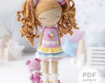 May include: Crochet doll with long brown hair in pigtails wearing a pink and yellow dress with a blue skirt and pink shoes. The doll is holding a pink crocheted teddy bear.