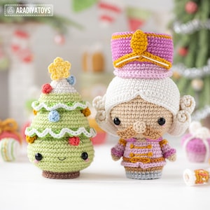 Op de afbeelding: Handgemaakte gehaakte kerstversieringen. Een lachende groene kerstboom met kleurrijke ballen en een ster, en een roze en gouden Notenkraker soldaat. Beide hebben zwarte knoopogen. Het Aradiva Toys-logo is zichtbaar.