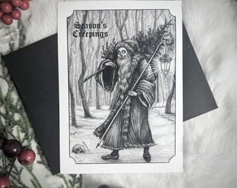 May include: A black and white illustration on a greeting card depicts a figure in a long coat and staff, carrying a Christmas tree in a snowy forest. The card reads "Season's Creepings". A black envelope is behind the card.