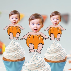 May include: Three cupcakes decorated with white frosting and blue sprinkles. Each cupcake has a pumpkin-shaped cutout with a baby's face inside. The baby is wearing a pumpkin costume.