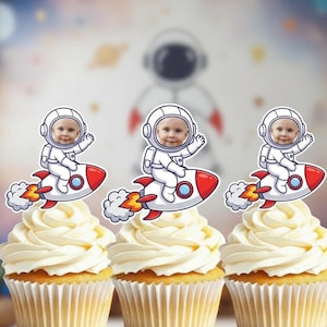 May include: Three cupcakes topped with white frosting and astronaut-themed toppers. Each topper features a baby's face in a white spacesuit riding a red and white rocket ship. The cupcakes are decorated for a space-themed party.