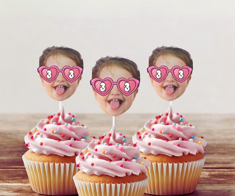 May include: Three cupcakes with pink frosting and colorful sprinkles. Each cupcake is topped with a photo of a child wearing heart-shaped glasses with the number 3. The cupcakes sit on a wooden surface.