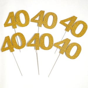 May include: Six gold glitter cake toppers, each with the number "40" printed on it. The toppers are on white sticks.