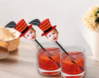 May include: Two red and gold ringmaster hats with faces of babies on top of black straws in two clear glasses filled with red liquid.