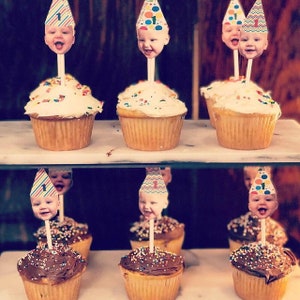 K&ouml;nnte beinhalten: Cupcakes mit wei&szlig;em und Schokoladen-Frosting, garniert mit bunten Streuseln. Jeder Cupcake hat ein Foto eines Babys mit einer Partyh&uuml;tchen mit der Zahl "1" auf einem St&auml;bchen. Die Cupcakes sind auf einem zweist&ouml;ckigen Regal angeordnet.