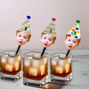 K&ouml;nnte beinhalten: Drei quadratische Gl&auml;ser gef&uuml;llt mit einer braunen Fl&uuml;ssigkeit und Eisw&uuml;rfeln. Jedes Glas hat einen schwarzen Strohhalm, der mit dem Gesicht eines Kindes mit einer Partyh&uuml;tchen bedeckt ist. Die H&uuml;te haben bunte Muster und die Zahl 1.