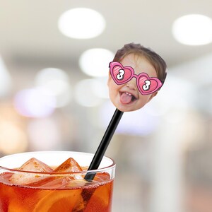 May include: A novelty drink stirrer featuring a child's face with heart-shaped glasses and the number 3. The stirrer is black and is placed in a glass of iced orange-colored liquid.