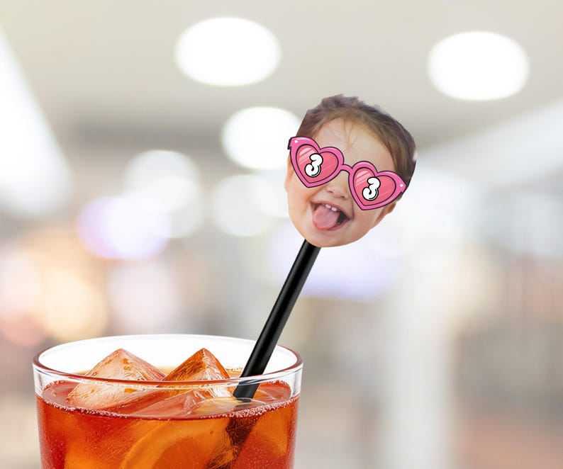 May include: A novelty drink stirrer featuring a child's face with heart-shaped glasses and the number 3. The stirrer is black and is placed in a glass of iced orange-colored liquid.