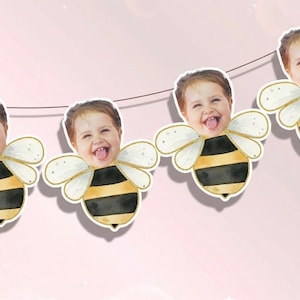 May include: A string of bee-shaped decorations with a child's face, black and yellow striped bodies, and white and gold wings. The garland is strung on a thin, dark string against a light pink background.