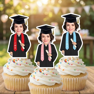 May include: Three cupcakes topped with white frosting and colorful sprinkles. Each cupcake has a graduation-themed cutout featuring a person in a cap and gown, holding a diploma. The cutouts have red, pink, and blue stoles.