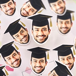 May include: A collection of graduation cap party decorations featuring a person's face on each cap. The caps are black with gold tassels. The faces are all smiling and have a variety of skin tones.