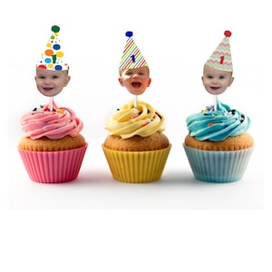 K&ouml;nnte beinhalten: Drei Cupcakes mit farbenfrohem Zuckerguss und dekorativen Toppern mit einem Babygesicht, das einen Partyhut tr&auml;gt. Die Cupcakes haben rosa, gelbes und blaues Frosting mit Streuseln. Jeder Cupcake sitzt in einer farblich passenden Papierform.