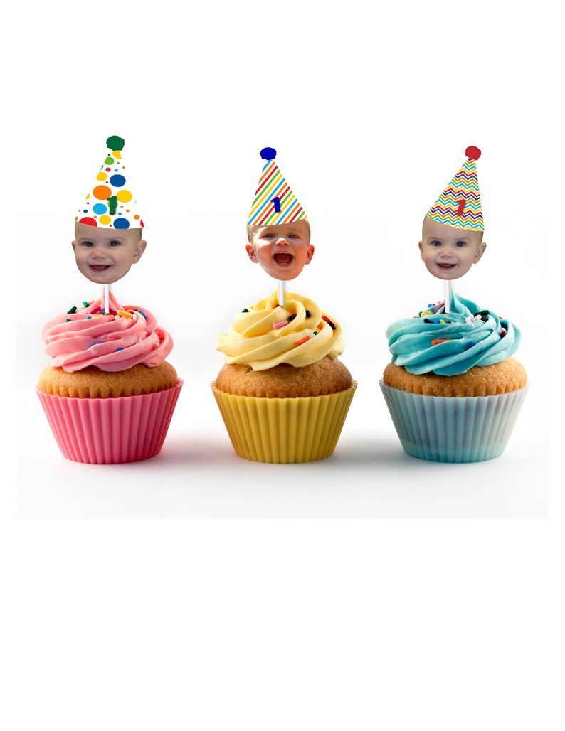 K&ouml;nnte beinhalten: Drei Cupcakes mit farbenfrohem Zuckerguss und dekorativen Toppern mit einem Babygesicht, das einen Partyhut tr&auml;gt. Die Cupcakes haben rosa, gelbes und blaues Frosting mit Streuseln. Jeder Cupcake sitzt in einer farblich passenden Papierform.
