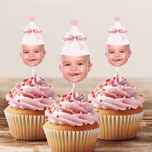 May include: Three pink and white cupcakes with pink frosting and colorful sprinkles topped with a baby face cupcake topper wearing a pink and white party hat.