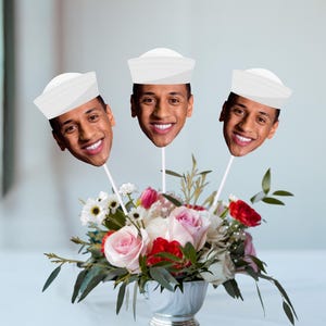 May include: A floral arrangement featuring three smiling faces wearing sailor hats on sticks. The flowers include pink and red roses, white daisies, and greenery in a silver vase. The background is a blurred white.