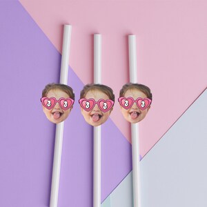 May include: Three white straws with attached images of a child's face wearing heart-shaped sunglasses with the number 3. The background is a geometric design of pink, purple, and white.