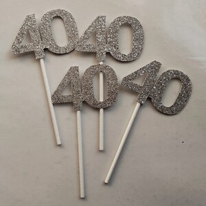 May include: Four silver glitter cake toppers shaped like the number 40. Each topper is attached to a white stick.