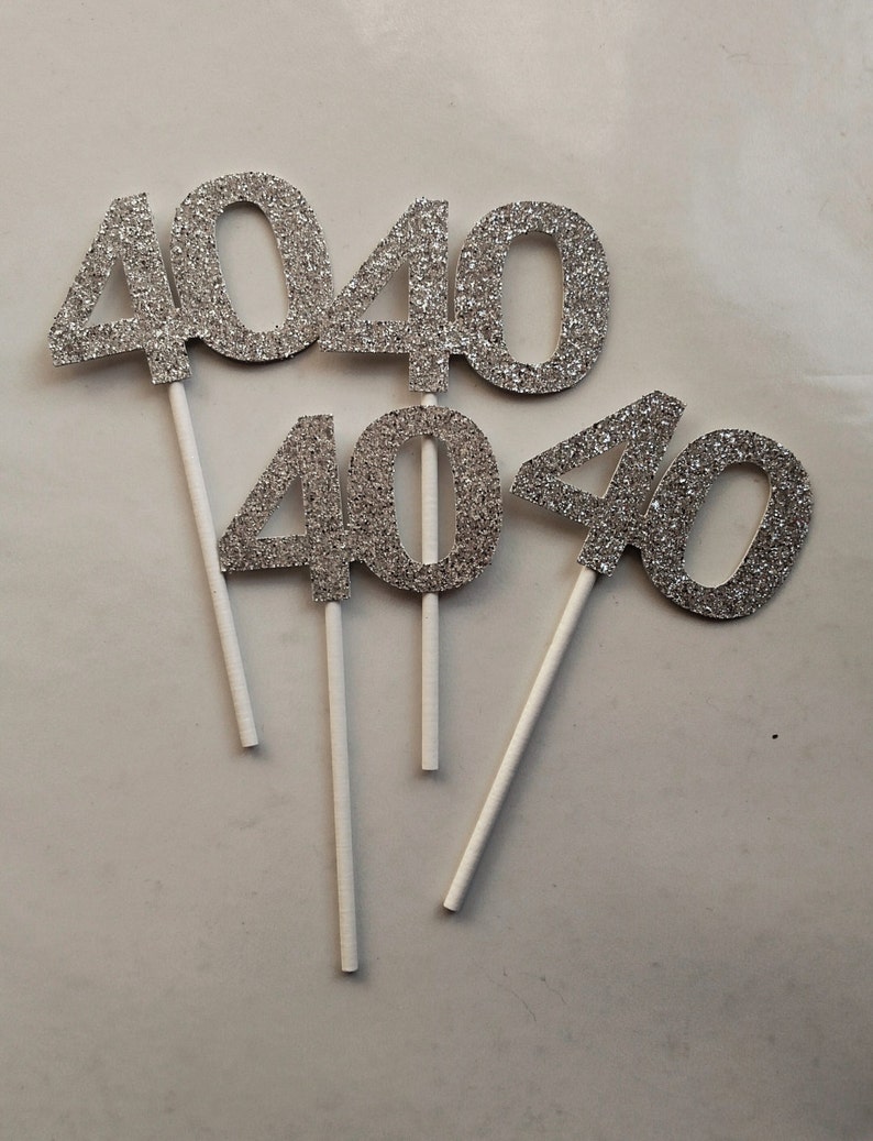 May include: Four silver glitter cake toppers shaped like the number 40. Each topper is attached to a white stick.
