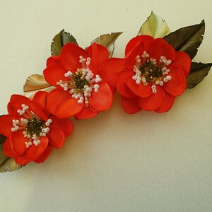 May include: Three orange fabric flowers with white and gold bead centers, arranged in a curved line with green fabric leaves.