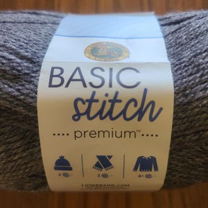 May include: A skein of gray yarn with a label that reads "BASIC stitch premium." The label also features icons for a hat, scarf, and sweater, with corresponding yarn weight recommendations. The Lion Brand logo is visible.
