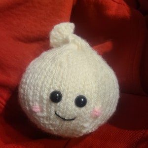 May include: A white knitted plush toy shaped like a garlic bulb with a smiling face, black eyes, and pink cheeks.